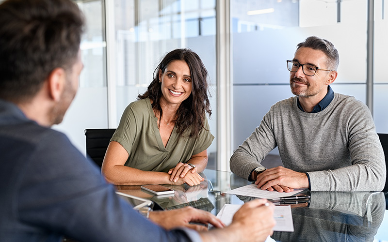 Three people sit at a table reviewing documents; two face the camera and smile while the third has his back to the viewer.