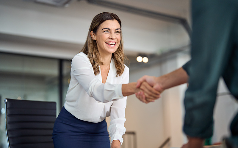 A woman at a desk facing the viewer shakes hands with a man who is mostly out of frame to the right.