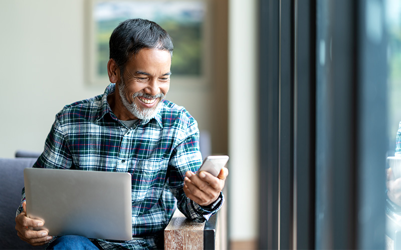 A man in a plaid shirt smiles while holding his laptop in one hand and looking at his phone in the other.