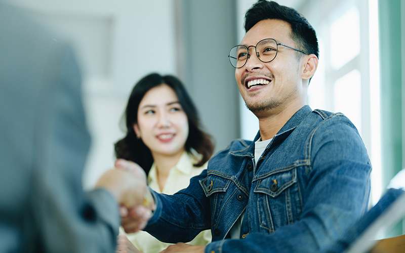 A smiling man shakes hands with a person who is mostly out of the frame while a third person looks on, also smiling.