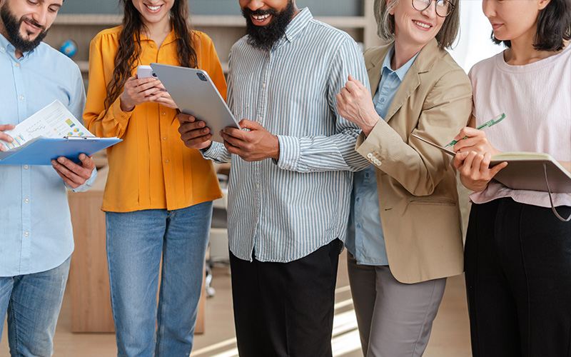 The man in the center of a group of people completes a form on a tablet. The others hold phones and project plans.