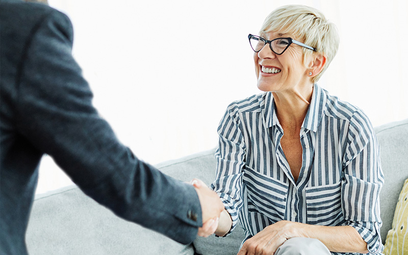 A woman sitting on a couch facing the viewer shakes hands with a man who is mostly out of frame to the left.